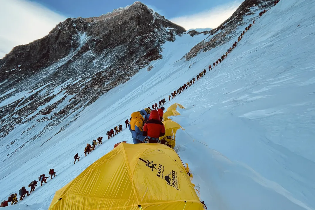 yellow tents in the foreground with a line of dozens of climbers in close proximity ascending up a steep snow-covered slope with mountain peaks in the background
